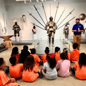 A group of children in orange t-shirts sits on the floor, observing a museum guide near a display of historical armor and weapons.