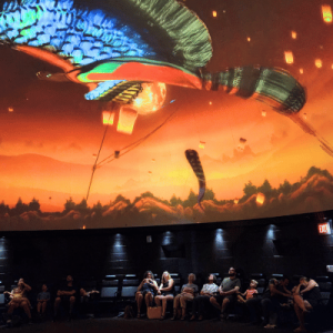 Visitors watch a vibrant digital display of a colorful serpent and lanterns on a dome ceiling in a planetarium.