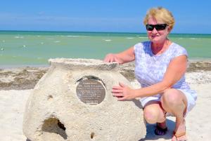 A woman in a blue shirt crouches beside a Memorial Reef, smiling at the camera. Behind her is a bright beach and sandy shore.