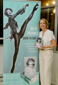 Author Juliet Prowse standing beside a banner of her dancing aunt with book "Juliet Prowse: Born to Dance". She's holding a book with the cover. The mood is celebratory.