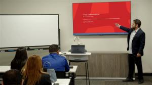 A man in a suit jacket stands at the front of a classroom, pointing at a presentation slide on a large screen that reads “The Constitution / La Constitución,” while several adult students sit at desks listening.