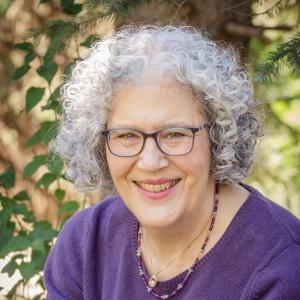 Author headshot of Susan Bruck, writer of "The DREAMER Path: A Journey to Rest, Renewal, and Your Authentic Self." She is smiling warmly while wearing glasses, a purple sweater, and a beaded necklace, set against a natural outdoor background with greenery