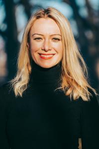 Barbara Murphy smiles at the camera in a black turtleneck, standing in natural light with a soft-focus background of trees. She has long blonde hair and a bright, confident expression.