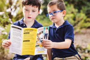 Two young boys, both light-skinned and brown haired, hold up a brochure in front of them to read about CO safety in school environments.