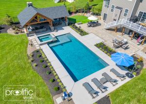 Aerial view of a modern backyard with a large rectangular swimming pool, attached hot tub, lounge chairs with a blue umbrella, outdoor covered seating area with bar and fireplace, and landscaped patio with dining space and putting green