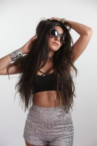 Farrah Mechael stands confidently with long hair flowing, wearing oversized round sunglasses, a black crop top, and silver sequin shorts. Her striking post reflects the bold energy of her cross-cultural pop sound.