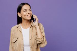 Woman smiling into the distance, in a light brown top, and with dark pulled back hair. She is on her mobile phone, with it held to her ear.