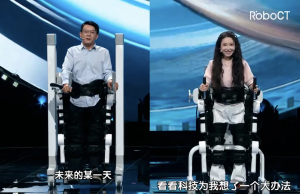 Two participants walking side-by-side in RoboCT rehabilitative exoskeletons on the CMG stage, therapists monitoring cadence, balance, and safety.
