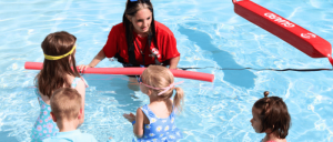A lifeguard in a red shirt teaches children in a swimming pool, using floatation devices to demonstrate water safety.