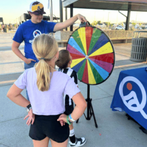 A young girl watches as a man spins a colorful prize wheel at an outdoor event, with another child beside her.