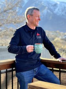 Man drinking coffee and wearing navy polo with the mountains in the background.