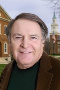 Headshot of Dr. S. Lee Funk, smiling in a brown blazer and green shirt, standing outdoors in front of a brick building with a clock tower and American flag.