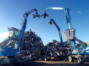 Two blue excavators with claw attachments working over large piles of scrap metal under a clear blue sky.