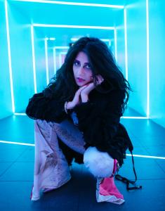 Farrah Mechael crouches in a glowing blue tunnel, framed by neon light bars. She wears flared pants with pink accents and a dark textured top, gazing directly into the camera with expressive intensity, echoing the cinematic mood of “Encore."