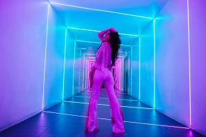 Farrah Mechael stands in a purple-and-blue neon-lit tunnel, posing with one arm raised behind her head. She wears lace flared pants and a sheer top, bathed in glowing light that mirrors the cinematic and futuristic aesthetic of “Encore.”