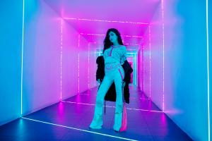 Farrah Mechael strikes a powerful stance in a neon-lit tunnel, wearing lace pants with hot pink lining and a matching fitted top, layered with a dark jacket draped behind her. The glowing blue and magenta lights amplify the strength and independent artist