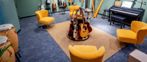 A cozy music room with yellow chairs surrounding a table of guitars, a piano, and percussion instruments, creating an inviting atmosphere.