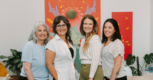 The Vervain Collective, a community apothecary and learning space dedicated to evidence-informed plant medicine, today announced that Good Apple has been appointed operating partner Vervain and Good Apple leaders standing together in the apothecary workspace, smiling before a vivid red floral mural and indoor plants