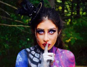 Farrah Mechael stands in a wooded setting with painted body art featuring skeleton details, glitter makeup, and silver accents. She wears violet contact lenses and a gothic headpiece, holding a finger to her lips as if to say “shh,” evoking mystery and th