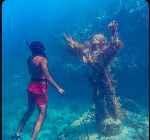 A snorkeler in red shorts swims respectfully beside the barnacle-covered bronze Christ of the Abyss statue, arms raised, in clear, blue shallow water with reef visible below.