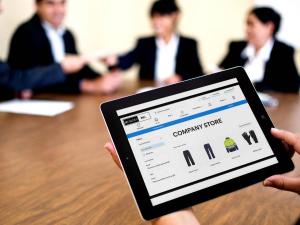 A person holds a tablet showing a custom company uniform store portal from Work Hard Dress Right, a Feury Image Group company, with work pants, safety gloves, and high-visibility gear displayed on the screen, while a team meets in the background, illustra