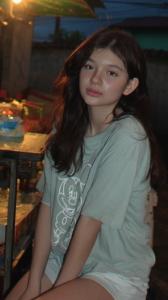 A hyper-realistic AI influencer, Mikaela Tala, sitting at a table in a dimly lit outdoor Filipino eatery at night.