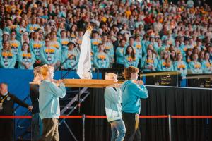 A group of young people walk in procession into the stadium, carrying a statue of Mary