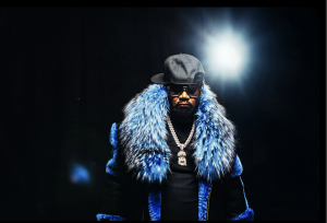 This is an image of HipHop Icon, GhostFace Killah. This is a vertical photo, GhostFace has on a royal blue fur jacket, black shades and a black hat. The background is black with a light flashing in the background