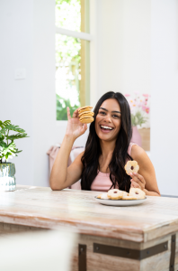 A smiling woman sitting at a table holding a stack of Cooee Cookies, with more cookies on a plate in front of her.