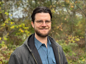 On Monday, Michigan 7th District Congressional candidate William Lawrence announced another 11 endorsements from current and former Mid-Michigan elected officials, bringing the total to 23 A photograph of a smiling man with glasses wearing a blue collared shirt and dark Carhartt coat. The background is fall woodlands.
