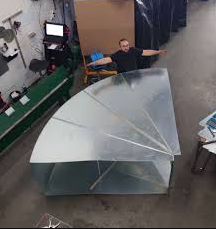 Person standing with arms outstretched behind a large, curved sheet metal duct section inside a workshop, showcasing the size and craftsmanship of the custom-fabricated piece.