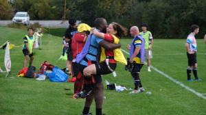 Coach Andrew Phillips lifts player Estelle McCalmont in celebration on the rugby field, both smiling after a game, surrounded by teammates and equipment bags.