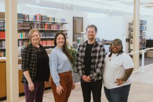 Photo of four smiling people standing side-by-side in a brightly lit space in front of a set of book shelves.