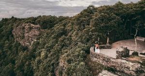A couple stands together on a cliffside terrace at Twickenham House with sweeping panoramic views of Blue Ridge Mountains overlooking a forested valley, with large natural rock formations in the foreground and clear blue sky above.