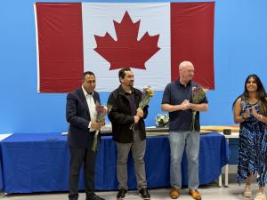 MPP Deepak Anand and two guests stand holding bouquets of flowers in front of a large Canadian flag during Zebra Robotics’ Annual Award Ceremony, as a presenter applauds in appreciation.