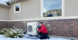 Photo of a modern Utah home exterior during winter, showing a professional installer (wearing a red collared shirt and black pants) installing an Amana heat pump near the side of the house. Includes energy savings icons like dollar signs and green leaves 