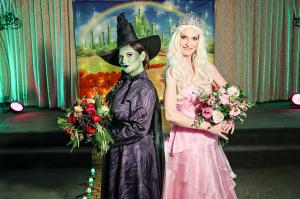 Famed for its theatrical, themed weddings, Viva Las Vegas Wedding Chapel launches its newest package catered to those whose love defies gravity A woman dressed as Elphaba in black with a green face and witches hat holds a large bouquet with dark flowers next to a woman dressed as Glinda in light pink with a crown holds a bouquet of light pinks and greens as they stand on a yellow brick road insid