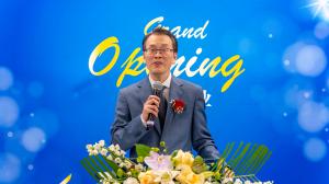 A man in a suit speaks into a microphone at a podium decorated with flowers during a Grand Opening Ceremony, with a blue background reading “Grand Opening.”