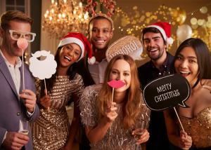 Diverse group of friends celebrating at a holiday party with festive decorations and photo booth props, holding "Merry Christmas" and "HO HO HO" signs, wearing Santa hats and sparkly attire, representing joyful authentic celebration.