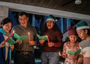 Multi-generational family of five caroling together indoors with candles and songbooks, wearing festive green elf hats and winter accessories.