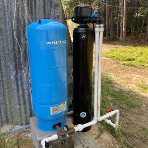 For many Mississippi homeowners who rely on private water wells for their daily needs, low water pressure might seem like a small inconvenience Outdoor well system with a blue WellXtrol pressure tank and a black filtration unit connected by PVC piping, installed beside a metal structure near a wooded area.