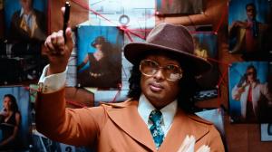 Ashwin Gane dressed as Inspector Gane, standing in front of a red-string evidence board and holding a pen while pointing upward in a noir-themed scene from the Flip Dat universe.