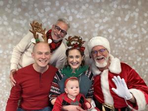 Two adults and a baby wearing reindeer and holiday accessories sit with Santa and another festive adult in front of a soft, sparkling backdrop, all smiling at a Sensory Santa event.