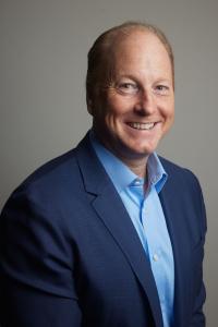 Tracy Maddux, beatBread interim CEO, smiling in a blue shirt with a blue jacket, against a grey background