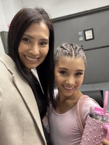 A woman and a young girl smile together in a close-up selfie taken indoors. The girl is dressed in light pink dancewear and holds a large pink beaded tumbler, while the woman wears a light-colored blazer. They are standing in front of a gray door backstag