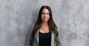 Portrait of financial coach and educator Hillary Seiler standing against a textured gray concrete wall. She’s wearing a gray jacket, black top and a delicate necklace, smiling confidently at the camera with long, styled brunette hair.