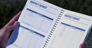 Close-up of the My Money Playbook monthly budget worksheets, showing sections for income, financial aid, college expenses and monthly spending. Hands hold the open spiral-bound financial planner outdoors in natural light.