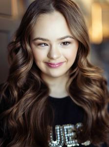 A close-up portrait of a smiling girl with long, softly curled brown hair and natural makeup. She is wearing a black top with rhinestone lettering and is looking slightly off to the side with a warm, relaxed expression. The background is softly blurred, g