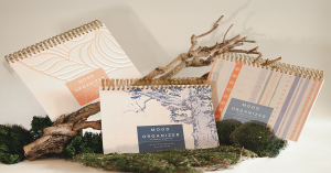 Three Mood Organizer wellbeing planners displayed with moss and driftwood for a natural, calming aesthetic Glimmery Moments®