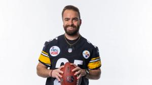 IgA Nephropathy Foundation and Steelers Back “13 for LIFE” as Hayden Shock Leads Awareness A man wearing a Pittsburgh Steelers jersey with a “Fan of the Year” patch smiles while holding a football against a plain white background.